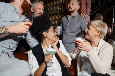 joyful multicultural colleagues drinking cocktails and talking in bar, after work relaxation clipart