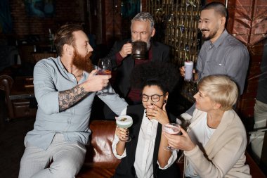 Sakallı, dövmeli bir adam işten sonra barda içki içip mutlu, etnik çeşitlilikte iş arkadaşlarıyla konuşuyor.