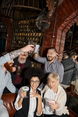 cheerful multicultural women looking at camera near bearded colleagues toasting in cocktail bar clipart