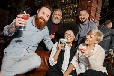 bearded tattooed man with cocktail looking at camera near multiethnic friends having fun in bar clipart