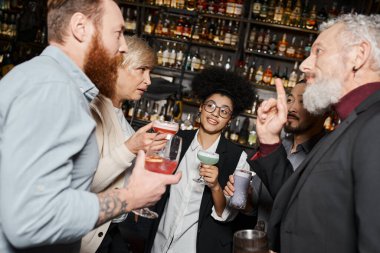 Fikir işareti gösteren sakallı bir adam. Barda kokteyl içen çok ırklı iş arkadaşlarıyla konuşuyor.