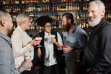 Sakallı, dövmeli adam, barda içki içip iş arkadaşlarının yanında gülümseyen Afrikalı Amerikalı bir kadınla konuşuyor.