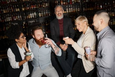 Çok ırklı iş arkadaşlarının yanında kameraya bakıp barda kokteyl içen neşeli sakallı adam.