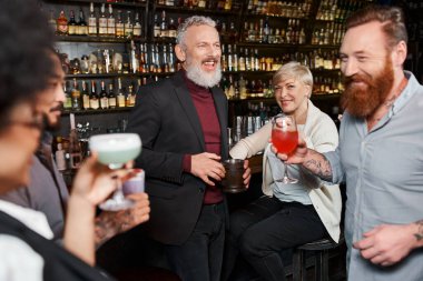Çok ırklı iş arkadaşlarının yanında gülen mutlu sakallı adam barda kokteyl tutuyor, parti zamanı.