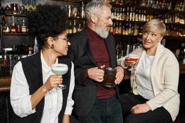 multiethnic women with cocktails glasses smiling during conversation with bearded colleague in bar clipart