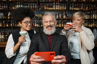 Mutlu sakallı adam, barda kokteyl bardakları olan çok kültürlü kadınlarla akıllı telefondan selfie çekiyor.