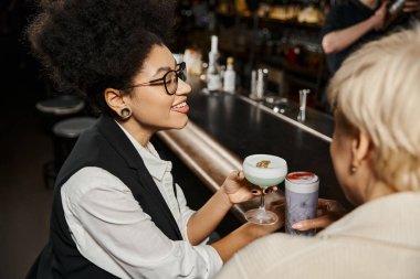 happy african american woman with blurred female colleague holding glasses with cocktails in bar clipart