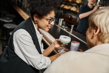 african american woman with cocktail glass smiling near female workmate in bar on blurred foreground clipart