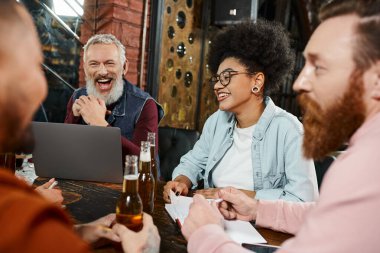 Heyecanlı sakallı adam dizüstü bilgisayarın yanında gülüyor ve çok ırklı iş arkadaşları barda bira şişeleriyle konuşuyor.