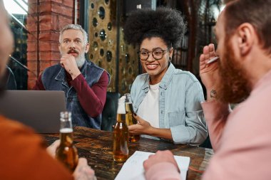 cheerful african american woman talking to multiethnic workmates during discussion on startup in pub clipart