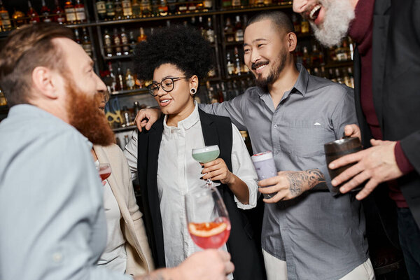 multicultural workmates holding glasses and smiling during conversation in cocktail bar after work
