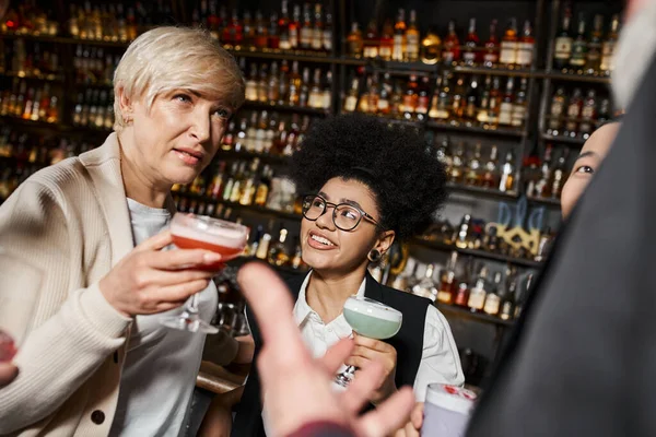 middle aged woman with cocktail glass talking to multicultural colleagues spending time in bar