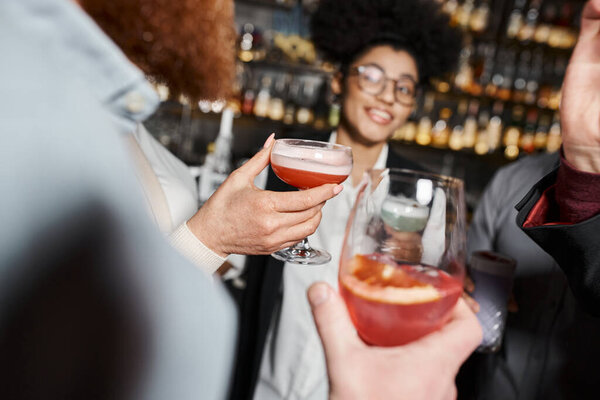 happy african american woman near colleagues with cocktail glasses in bar, blurred background