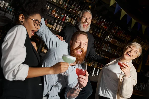 overjoyed multiethnic colleagues with cocktails laughing in bar, diverse team having fun after work