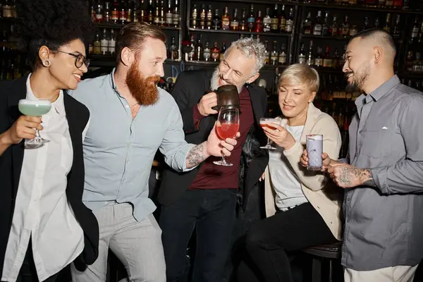 tattooed bearded man holding glass near cheerful multiethnic colleagues in bar, after work leisure