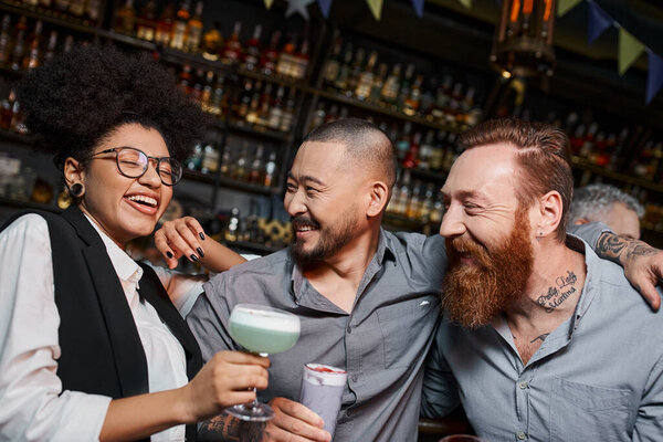 excited african american woman laughing near multiethnic bearded colleagues in bar, party time
