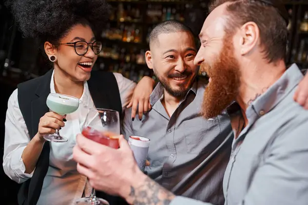 cheerful multiethnic work friends with glasses hugging and laughing in cocktail bar after work