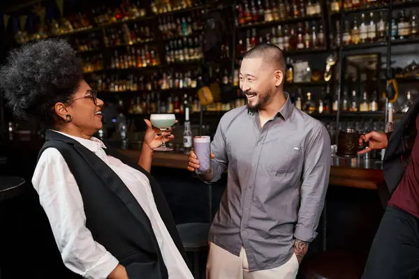 cheerful asian man and african american woman with drinks smiling at each other in cocktail bar