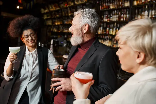 stock image bearded middle aged man smiling near multiethnic women with cocktails, colleagues resting in bar
