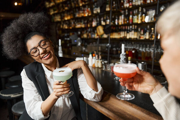 cheerful african american woman holding cocktail glass near female colleague on blurred foreground