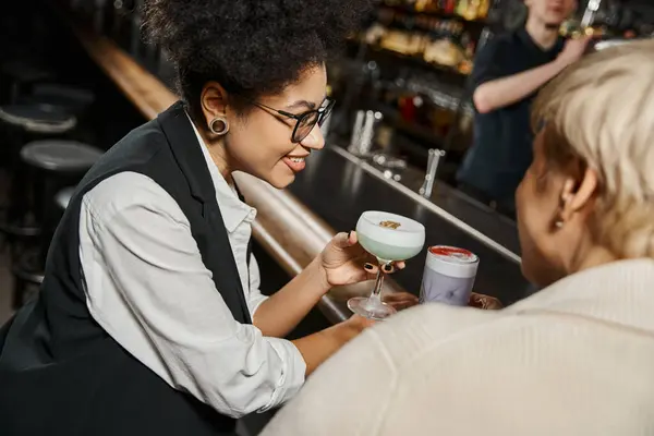 african american woman with cocktail glass smiling near female workmate in bar on blurred foreground