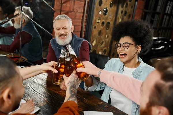 cheerful multiethnic work friends clinking beer bottles while spending after work time in bar