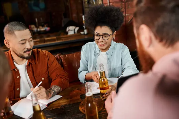 happy african american talking to multiethnic colleagues sitting near beer bottles in pub after work