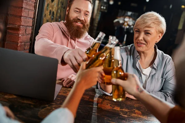 cheerful bearded man and middle aged woman clinking beer bottles with colleagues near laptop in pub