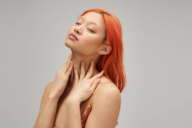 beautiful asian woman with dyed red hair and closed eyes touching her neck on grey background clipart