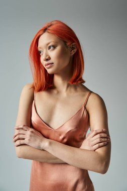 Kızıl saçlı gülümseyen genç Asyalı kadın pastel bir elbise giymiş gri arka planda, başka tarafa bakıyor.