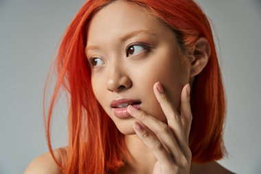 Asyalı güzel, kızıl saçlı ve doğal makyajlı genç bir kadın uzağa bakıyor ve yanaklarına dokunuyor.