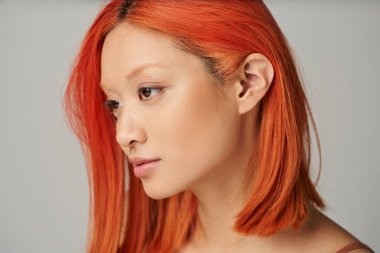 Gri arkaplanda poz veren mükemmel ciltli ve kızıl saçlı genç Asyalı kadının portresi.