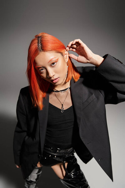 стильная азиатская женщина позирует в черном блейзере и регулирует рыжие волосы на сером фоне, смелый стиль