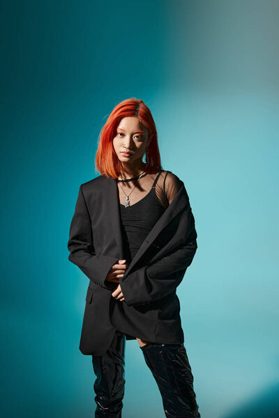 молодая азиатская женщина с окрашенными волосами позирует в огромных пиджаках и латексных сапогах на синем фоне