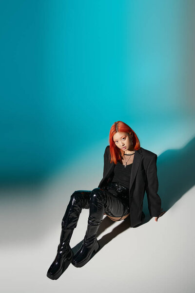 high angle, asian woman with nose piercing sitting in oversized blazer and black latex boots on blue