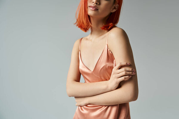 cropped view of elegant and young woman with red hair in pastel slip dress posing on grey background