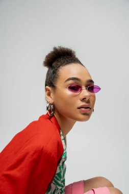 Genç Afro-Amerikan modelinin portresi şık giyinmiş ve gri arka planda pembe güneş gözlüğü takıyor.