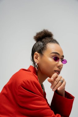 portrait of young african american model in stylish attire and pink sunglasses on grey backdrop clipart