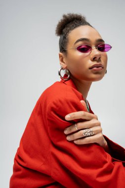 Güzel Afro-Amerikan modelinin portresi şık giyinmiş ve gri arka planda güneş gözlüğü takıyor.