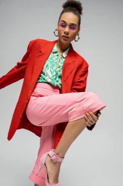 Gri arka planda, dinamik pozda, kalın giyimli ve güneş gözlüklü modacı Afro-Amerikan modeli