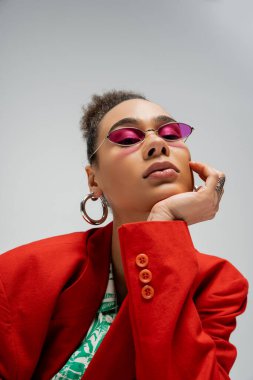 Pembe güneş gözlüklü ve takılı Afro-Amerikan model gri arka planda kameraya bakıyor.