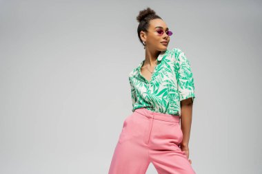pleased african american woman in pink sunglasses and stylish attire posing on grey backdrop clipart
