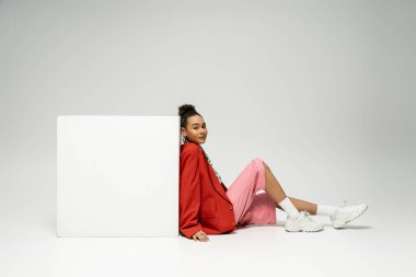 pretty african american woman in vibrant outfit sitting near cube on grey backdrop, look at camera clipart