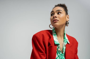 african american woman in stylish red blazer and hoop earrings looking away on grey backdrop clipart