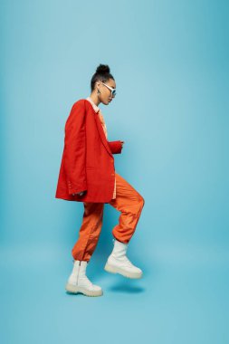 Uzun boylu, göze çarpan, güneş gözlüklü, parlak kıyafetli, mavi üzerinde yürüyen Afro-Amerikan kadın.