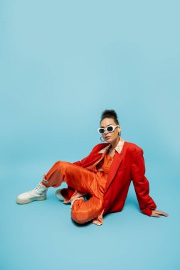 Kişisel stil, şık Afro-Amerikan modeli canlı kıyafetiyle mavi arka planda oturuyor.