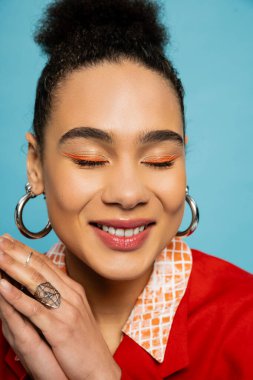 Mavi arka planda gülümseyen gümüş aksesuarlı mutlu Afro-Amerikan modelinin portresi