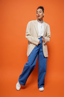 Renkli bej ceketli ve turuncu arka planda mavi pantolonlu Afro-Amerikan modeli.