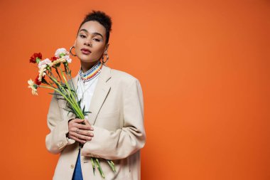 Güzel Afro-Amerikan modeli şık şehir kıyafetleri içinde çiçeklerle poz veriyor, turuncu arka plan