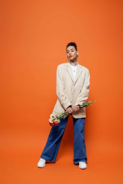 stylish fashion model in blue pants and beige blazer holding flowers in hands on orange backdrop clipart